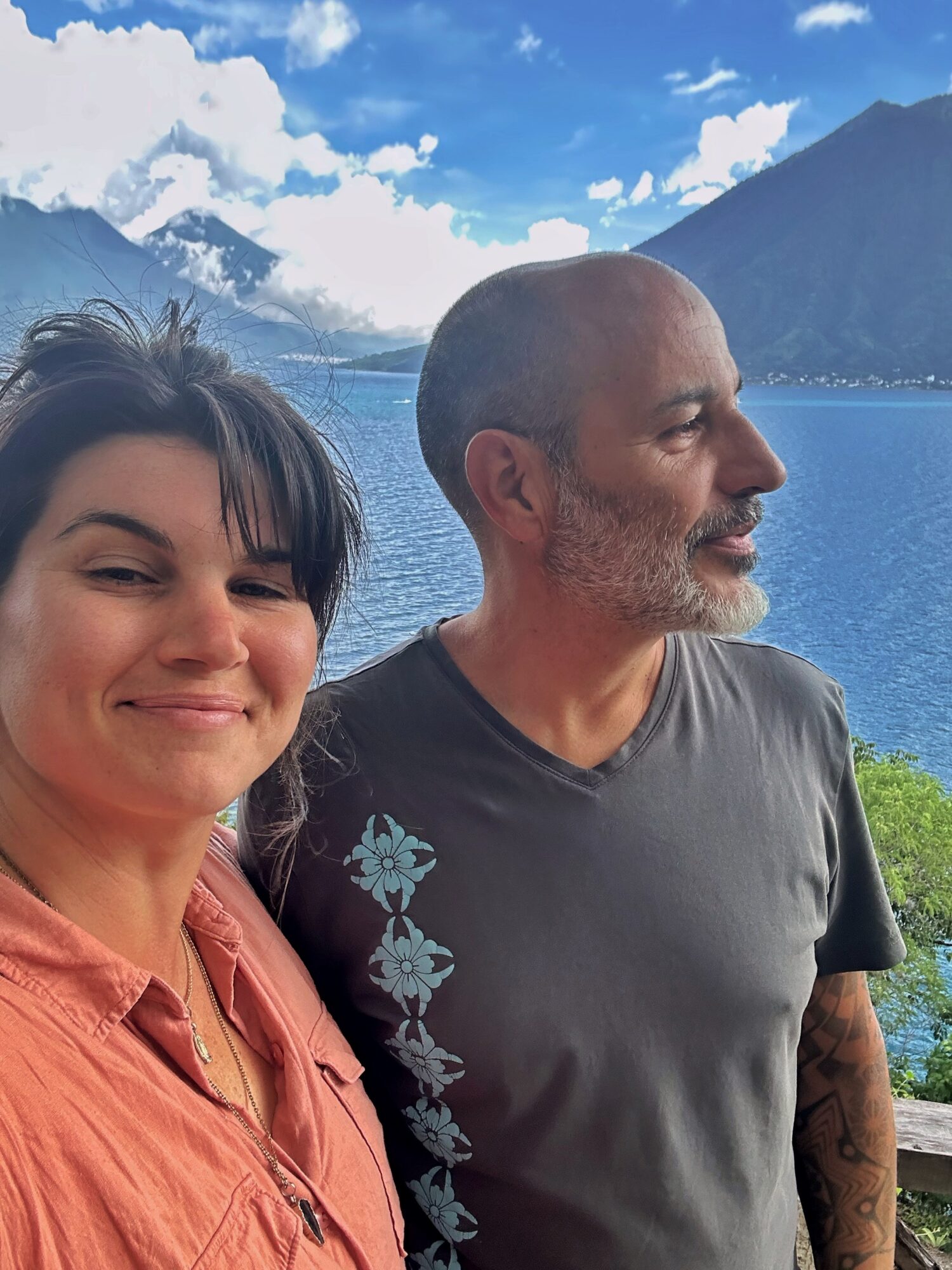 Two people pose beside a blue lake, with mountains and clouds in the background.