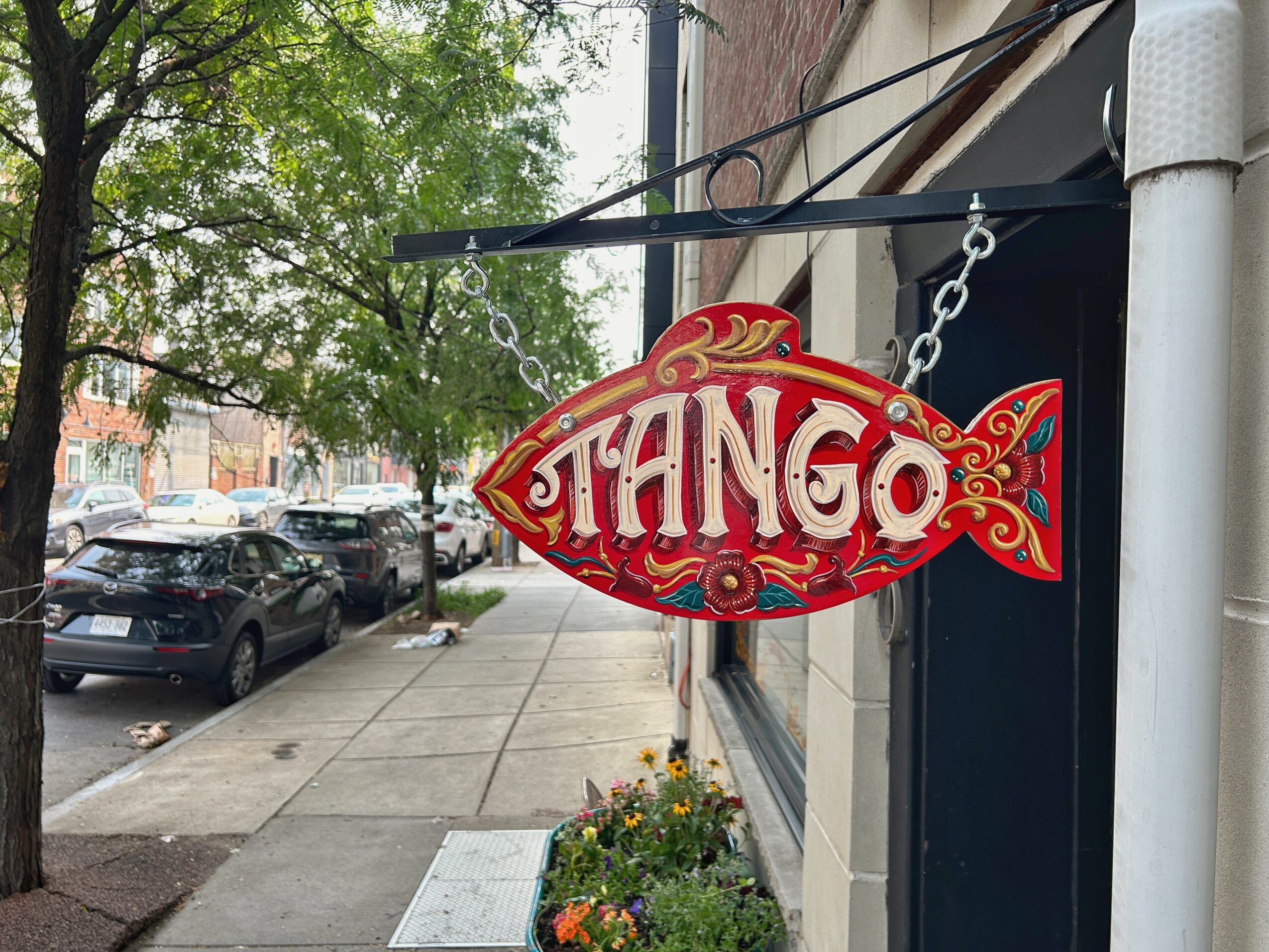 Ornate red and gold TANGO sign hangs above a sidewalk beside parked cars and a flower planter.