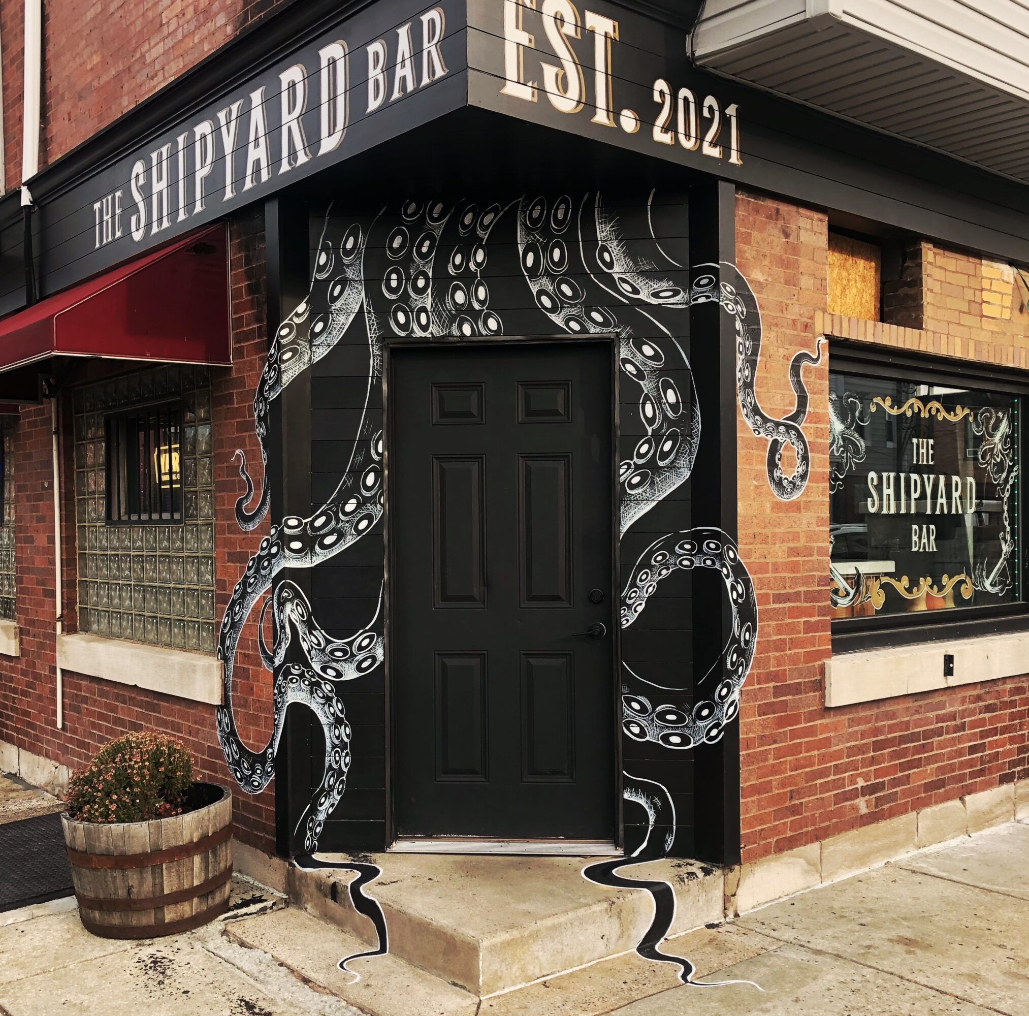 Brick bar entrance with a black door, octopus mural, barrel planter, and signage reading “The Shipyard Bar.”