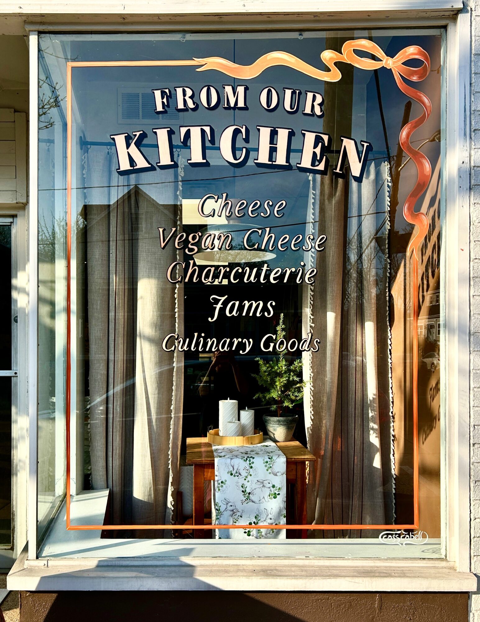Shop window sign reads “From Our Kitchen,” with cheese, vegan cheese, charcuterie, jams, and culinary goods listed.