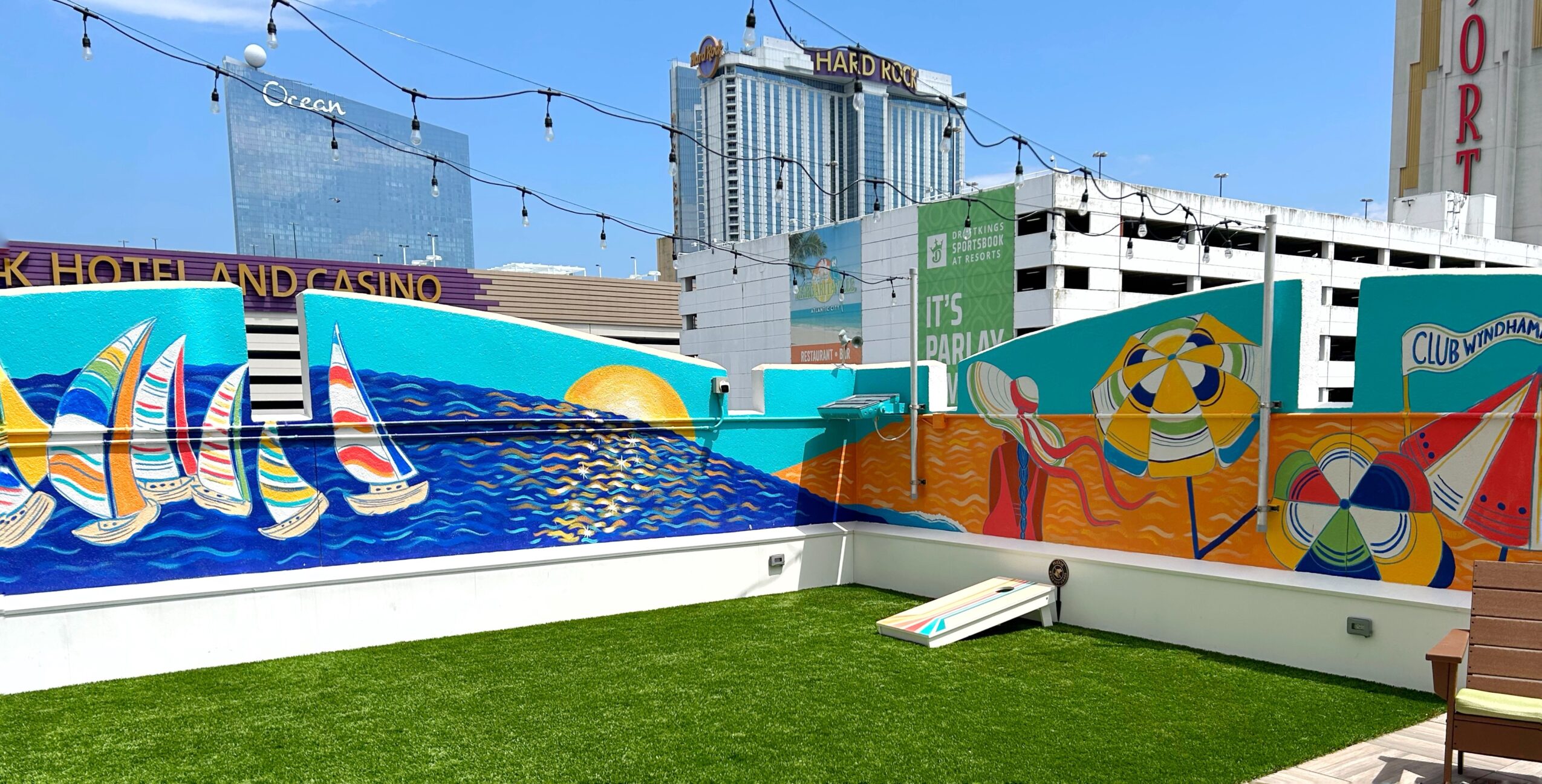 Artificial grass courtyard bordered by colorful mural walls, with hotel buildings and string lights in the background.