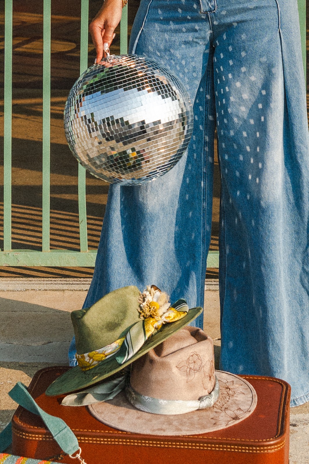A hand holds a mirrored disco ball beside wide-leg jeans, above a decorated hat resting on a suitcase.