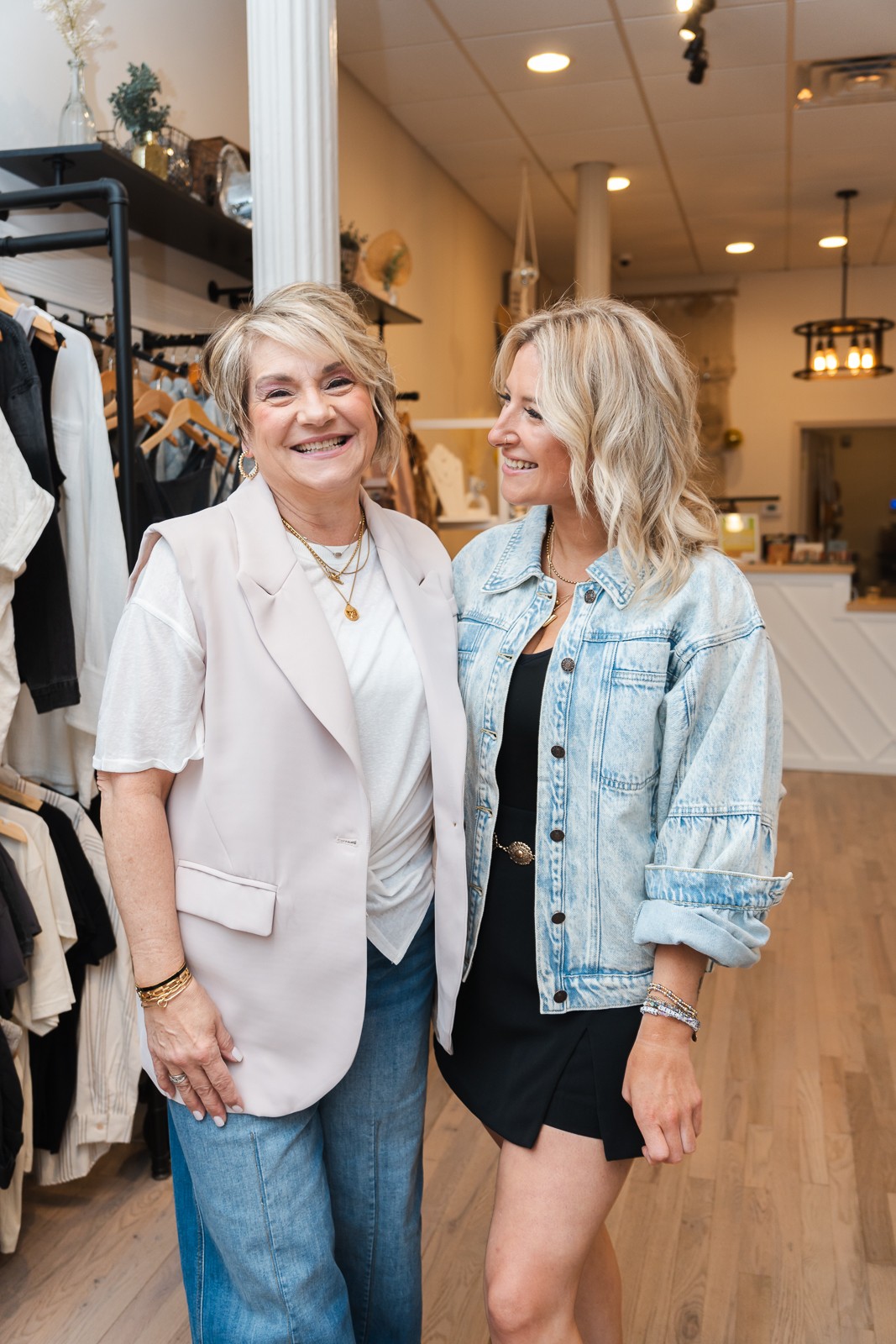 Two people stand together in a clothing boutique, with clothing racks and shelves behind them.