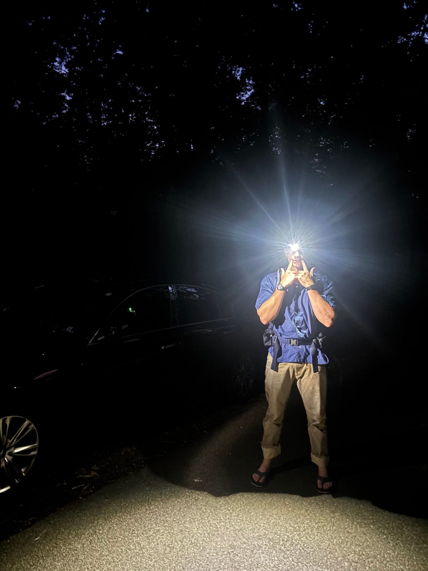 A person stands on a path at night, shining a bright light toward the camera beside a parked car.