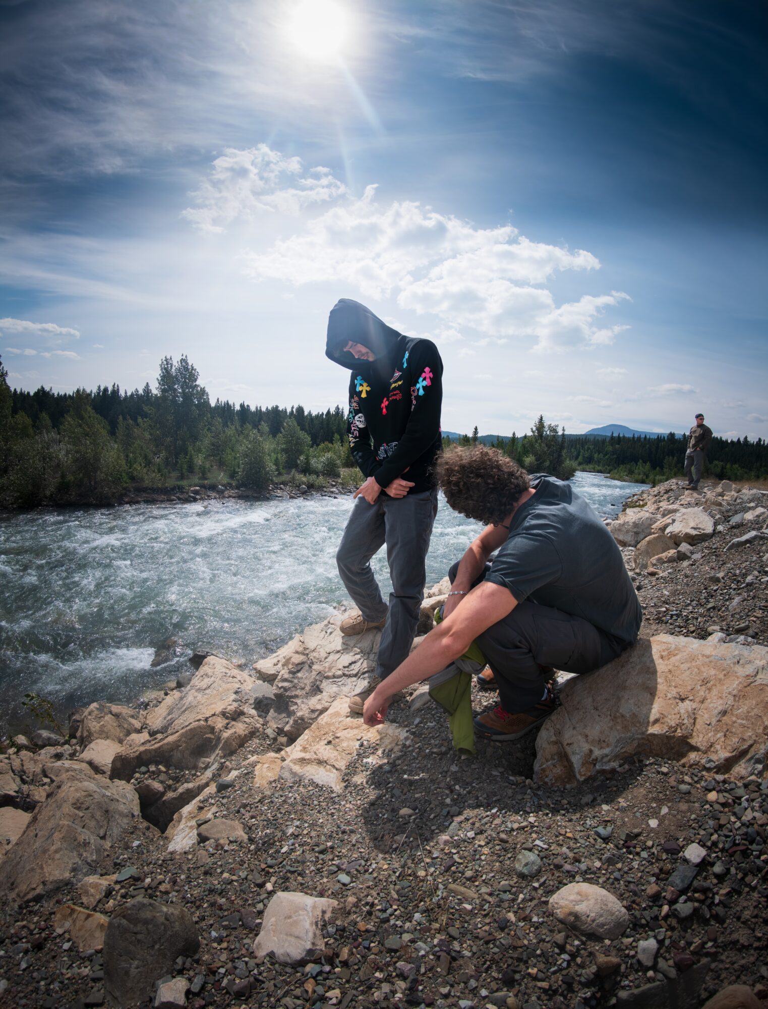 Two people crouch on a rocky riverbank beside flowing water, with trees and a bright sky in the background.