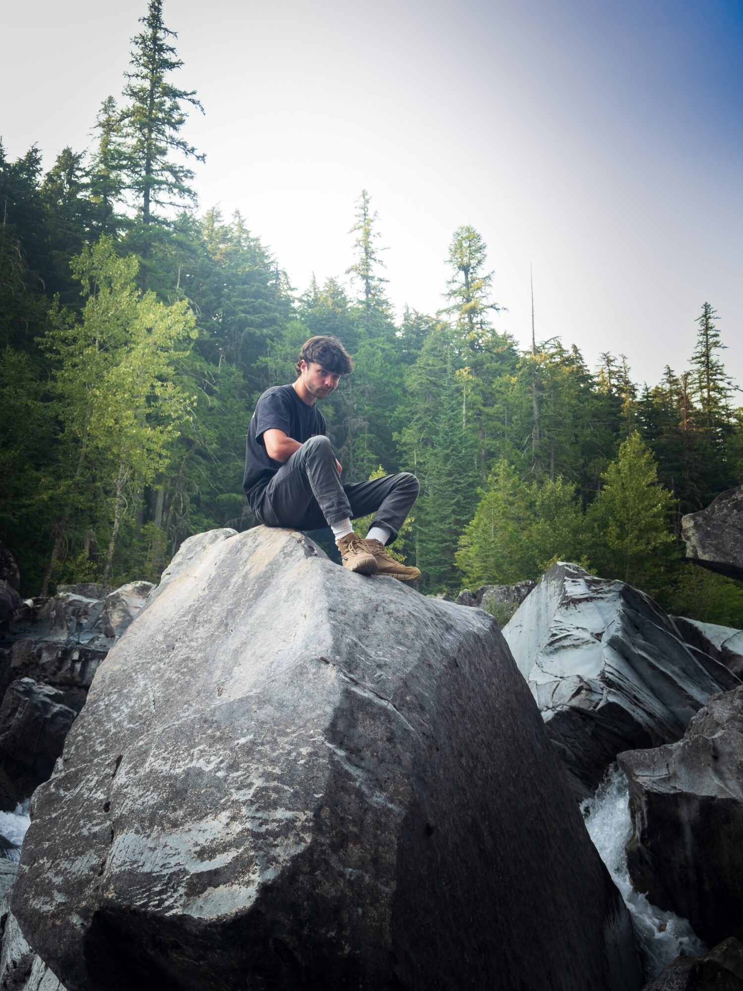 A person sits on a large boulder beside a rocky stream, surrounded by evergreen trees.