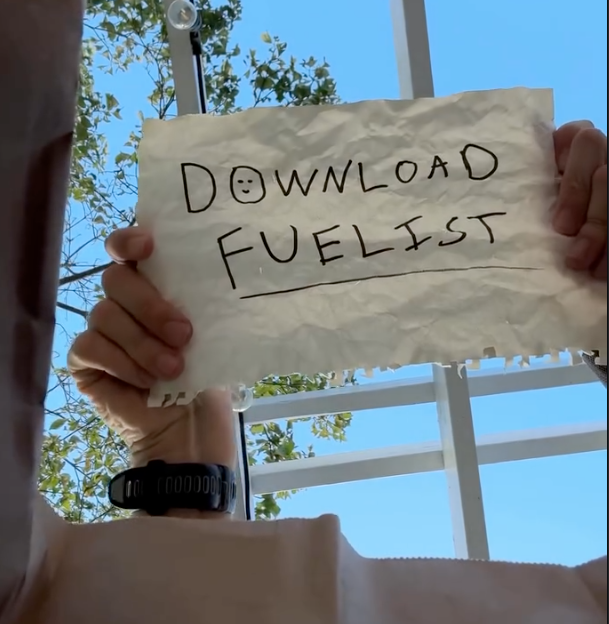 A person holds a handwritten paper sign reading “Download Fuelist” against a bright blue sky and leafy branches.